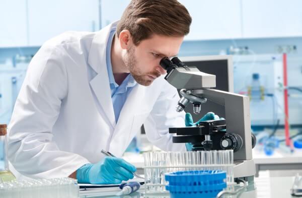 Atendimento laboratorial - homem branco de barba, com janelo, olhando um microscópio em um laboratório.