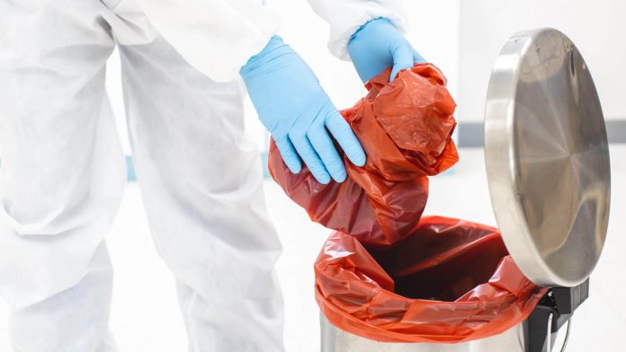 Profissional com EPI inserindo saco em recipiente, realizando descarte de resíduos laboratoriais.
