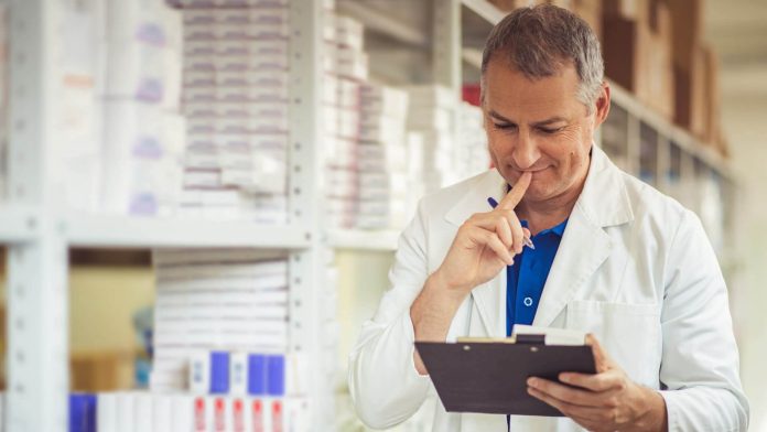 Homem branco, grisalho, com jaleco branco olhando para prancheta, realizando controle de estoque no laboratório.